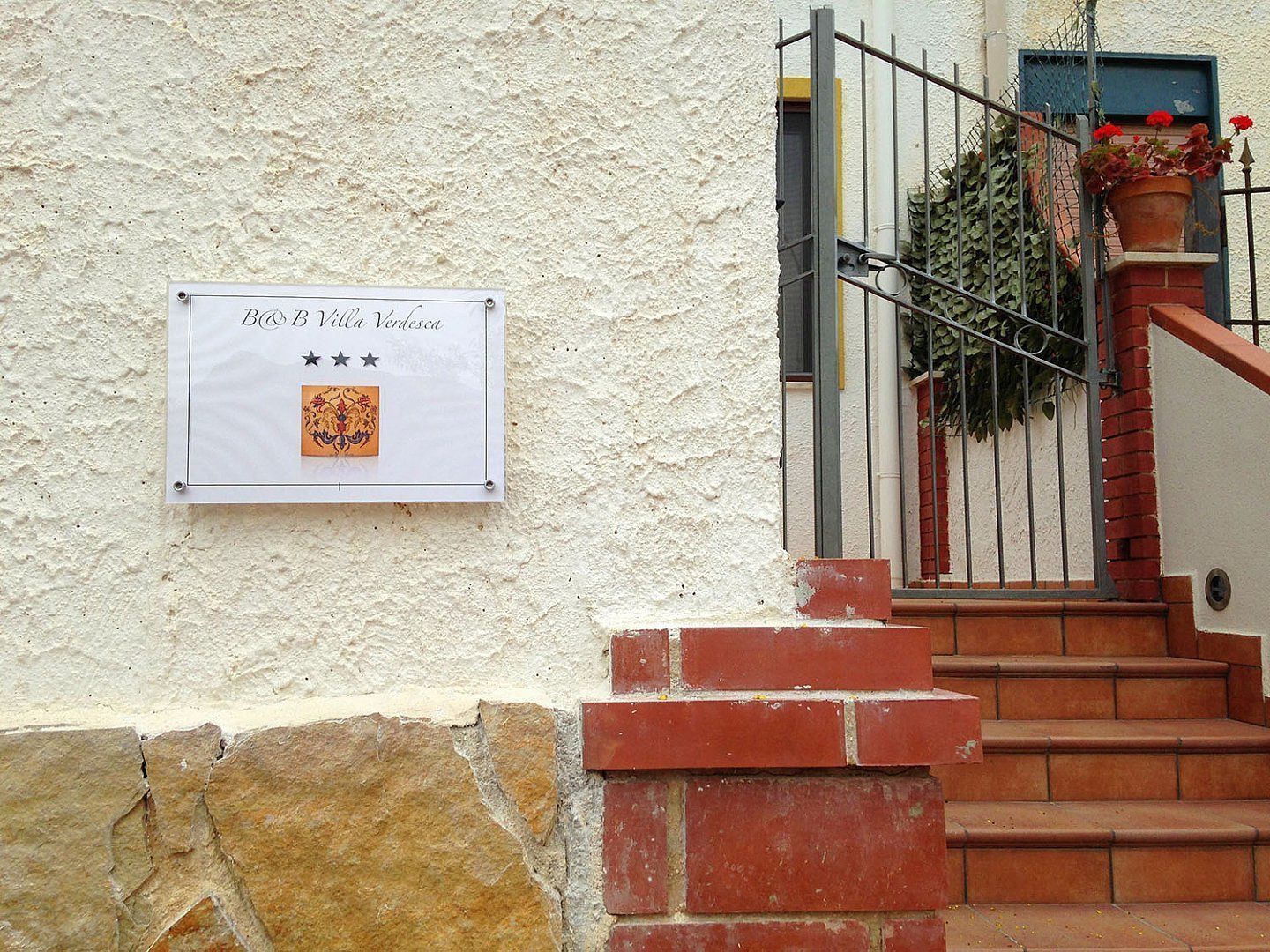 A sign with text and a coat of arms, a staircase with red bricks, and a metal railing with potted plants.