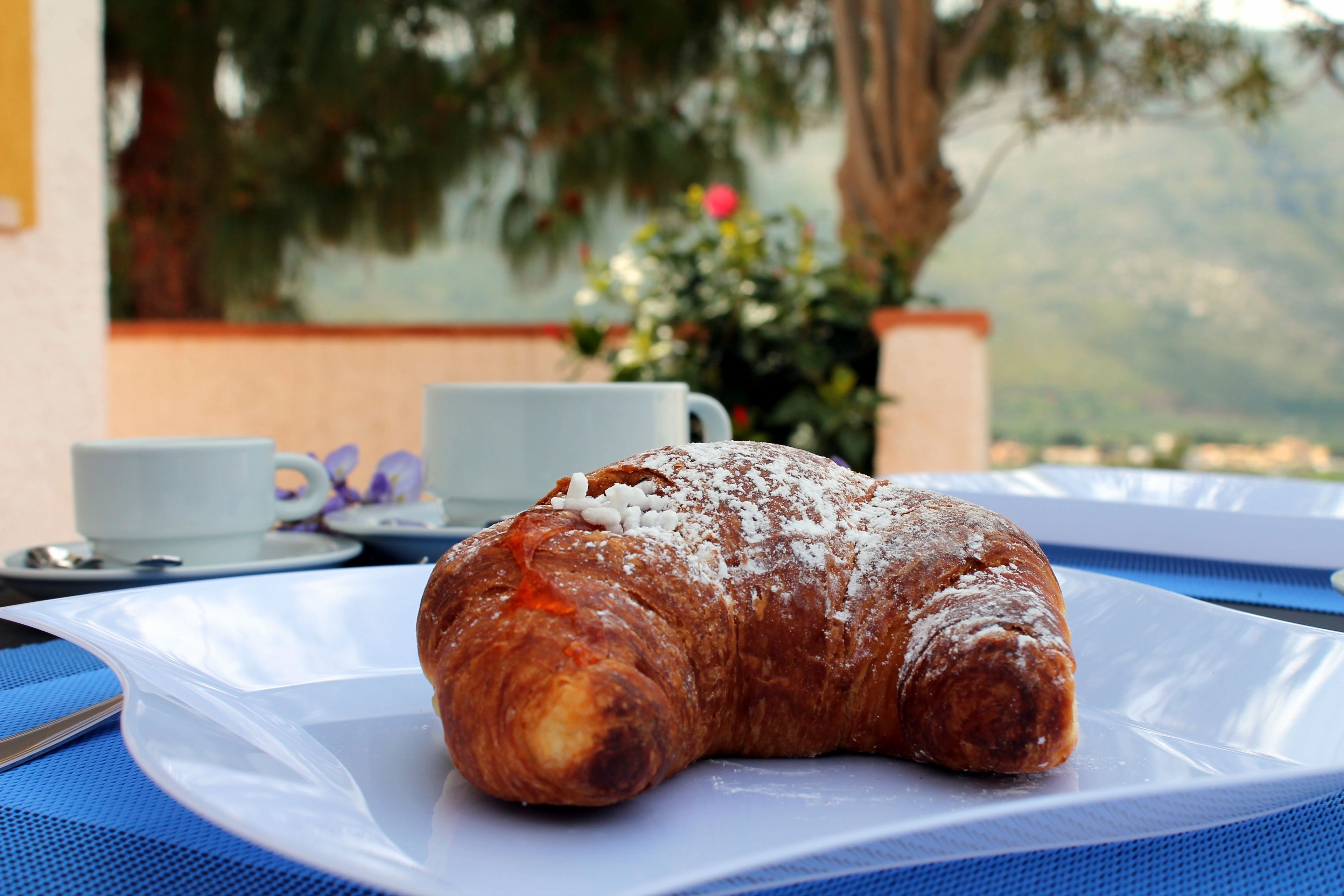A croissant on a white plate with a blue tablecloth, a white cup, and a small plate with a spoon on a table.