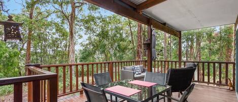 A covered outdoor seating area with a table and chairs, surrounded by a wooden deck and overlooking a forested area.