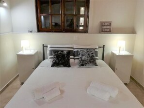 A neatly made bed with white linens and two pillows, two bedside tables with lamps, a window with a view, and a sign on the wall.