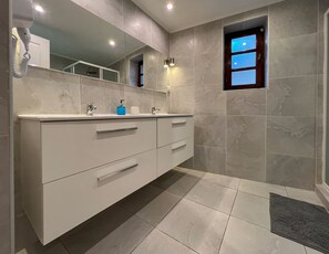 A modern bathroom with a large mirror, a vanity with drawers, a shower area, and a small window.