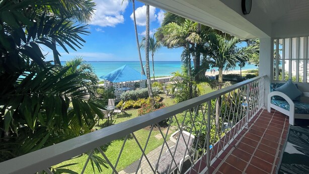 A balcony with a view of the ocean, surrounded by palm trees and a patio area with chairs and a table. - Beautiful One Bedroom Ocean View Gem (Spring Garden)