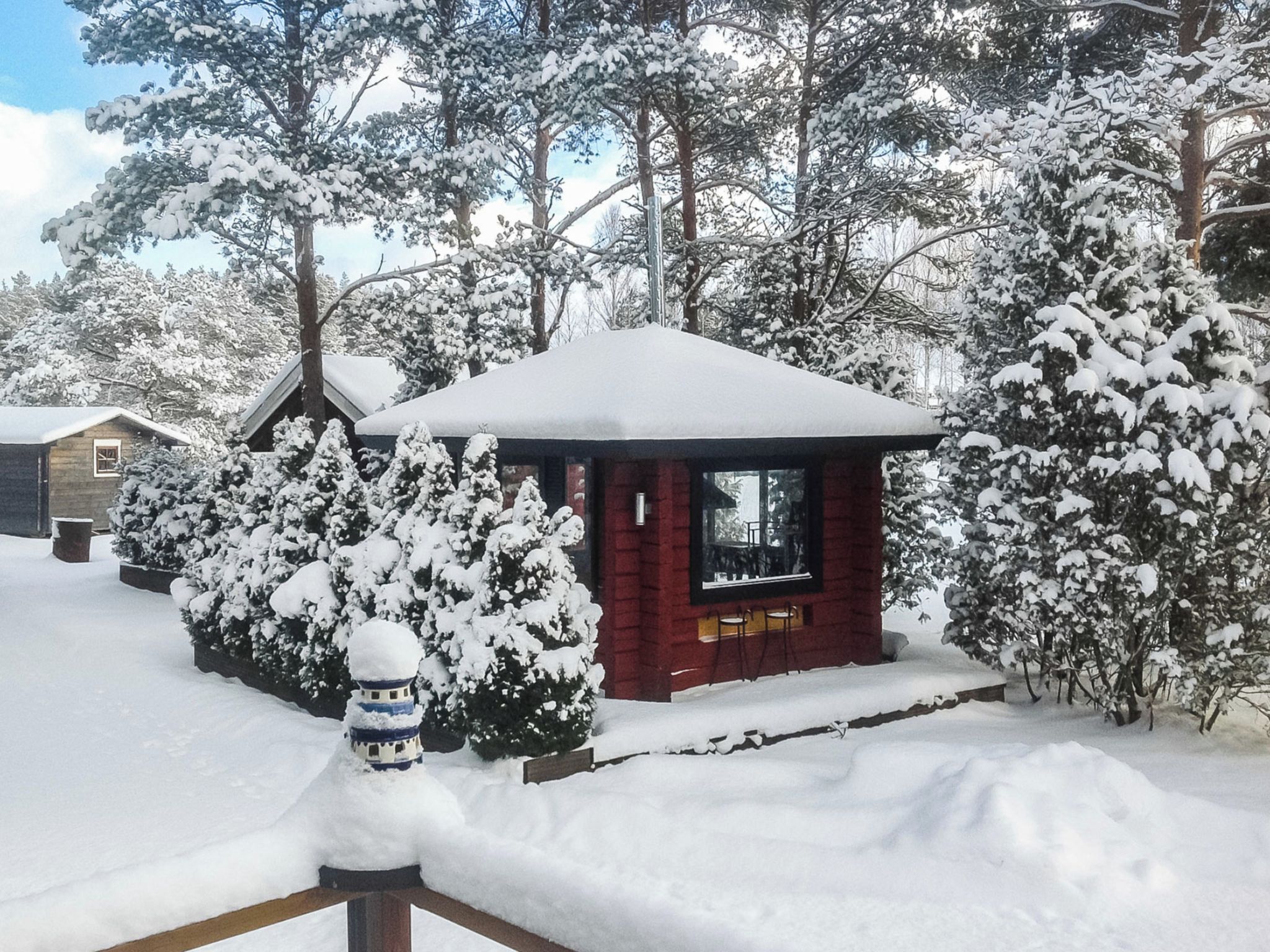 A small cabin with a snow-covered roof, surrounded by snow-laden trees and bushes.