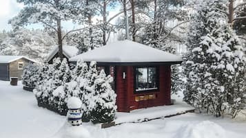 A small cabin with a snow-covered roof, surrounded by snow-laden trees and bushes.