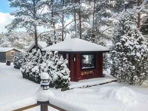 Una cabaña pequeña con techo cubierto de nieve, rodeada de árboles y arbustos cargados de nieve.