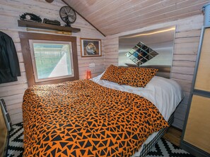 Una habitación con cama, ventana con persianas, ventilador y espejo en la pared.