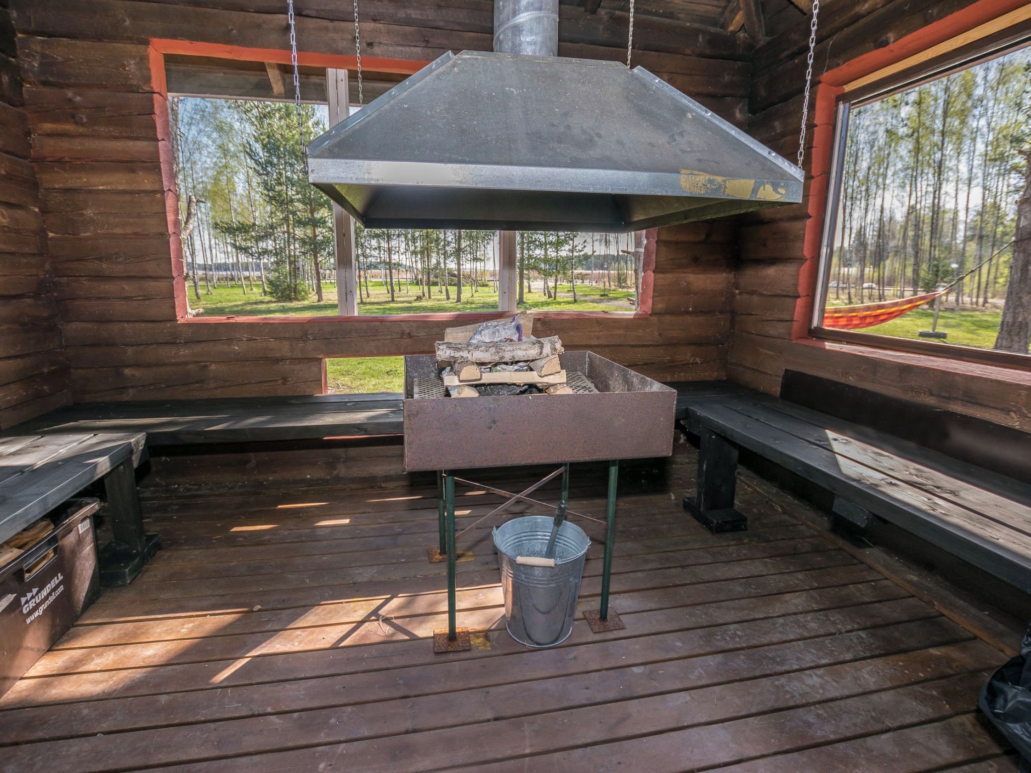 L’intérieur d’un chalet rustique, avec un barbecue en métal, des bancs en bois et une vue sur une forêt, visible à travers de grandes fenêtres.