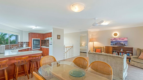 A modern kitchen with wooden cabinets, a dining area with a glass table, and a living room with a sofa and a TV.