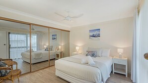 A hotel room with a large mirror, a bed, two bedside tables with lamps, a ceiling fan, and a view of the outdoors. - Ocean Waves - Downtown Oceanfront (Lennox Head)