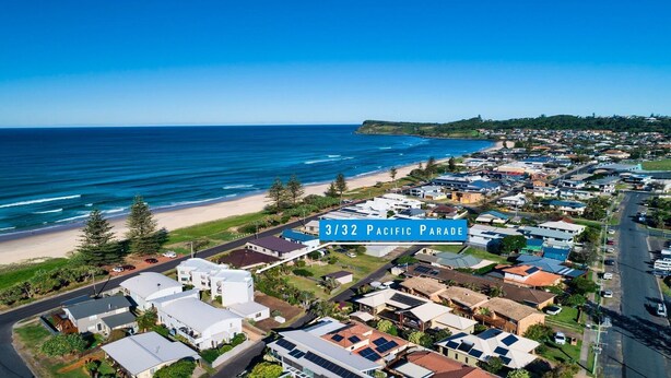 An aerial view of a coastal residential area with houses, a beach, and a sign indicating "3/32 Pacific Parade". - Ocean Waves - Downtown Oceanfront (Lennox Head)