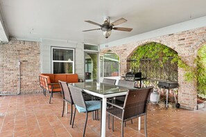 A patio with a glass table and chairs, a ceiling fan, brick walls, and a brick archway with climbing plants. - Private Retreat w/ Pool, Patio, BBQ! 6BR 3BA Gem (Houston)