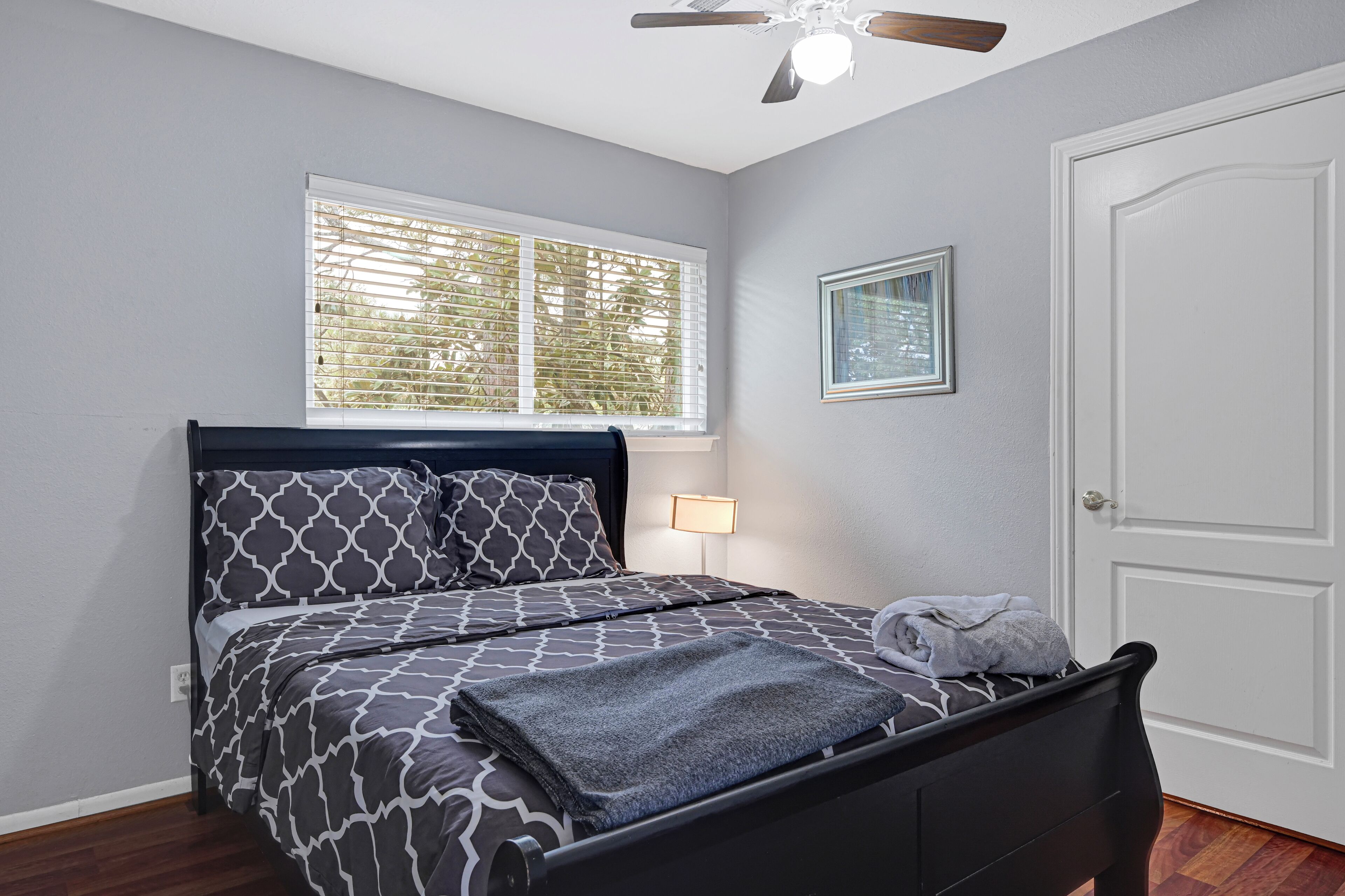 A neatly made bed with patterned bedding, a bedside lamp, a window with blinds, and a door.