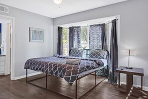 A bedroom with a bed, a bedside table, a window with curtains, and a door. - Private Retreat w/ Pool, Patio, BBQ! 6BR 3BA Gem (Houston)