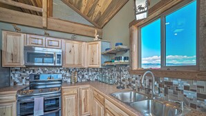 A kitchen with wooden cabinets, a stainless steel sink, and a large window offering a view of the sky. - Day Trip to Yellowstone NP! Mtn-View Cabin (Livingston)