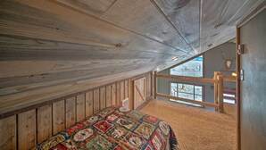 A loft bedroom with a wooden ceiling, a large bed with a colorful quilt, and a view of a snowy landscape through a window. - Day Trip to Yellowstone NP! Mtn-View Cabin (Livingston)