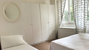 Un dormitorio con dos camas, un armario, un espejo, una ventana con cortinas y una pequeña mesa.