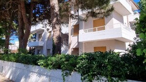 Un edificio residencial de varios pisos con balcones, rodeado de árboles y un muro blanco.