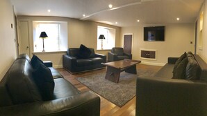 A living room with a sofa, armchair, and coffee table, a TV mounted on the wall, and a window with blinds. - No14 in the heart of St Andrews (St Andrews)