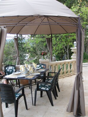 An outdoor dining area with a covered table and chairs, surrounded by a stone balustrade and greenery. - Provencal farmhouse in natural stones in a wooded hill with swimming pool. (Carcès)