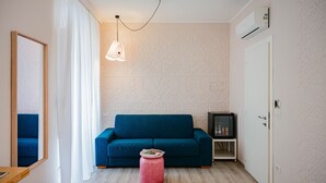 A modern living room with a blue sofa, a pink ottoman, a hanging light fixture, and a small entertainment unit. - A' Coffa (Taormina)