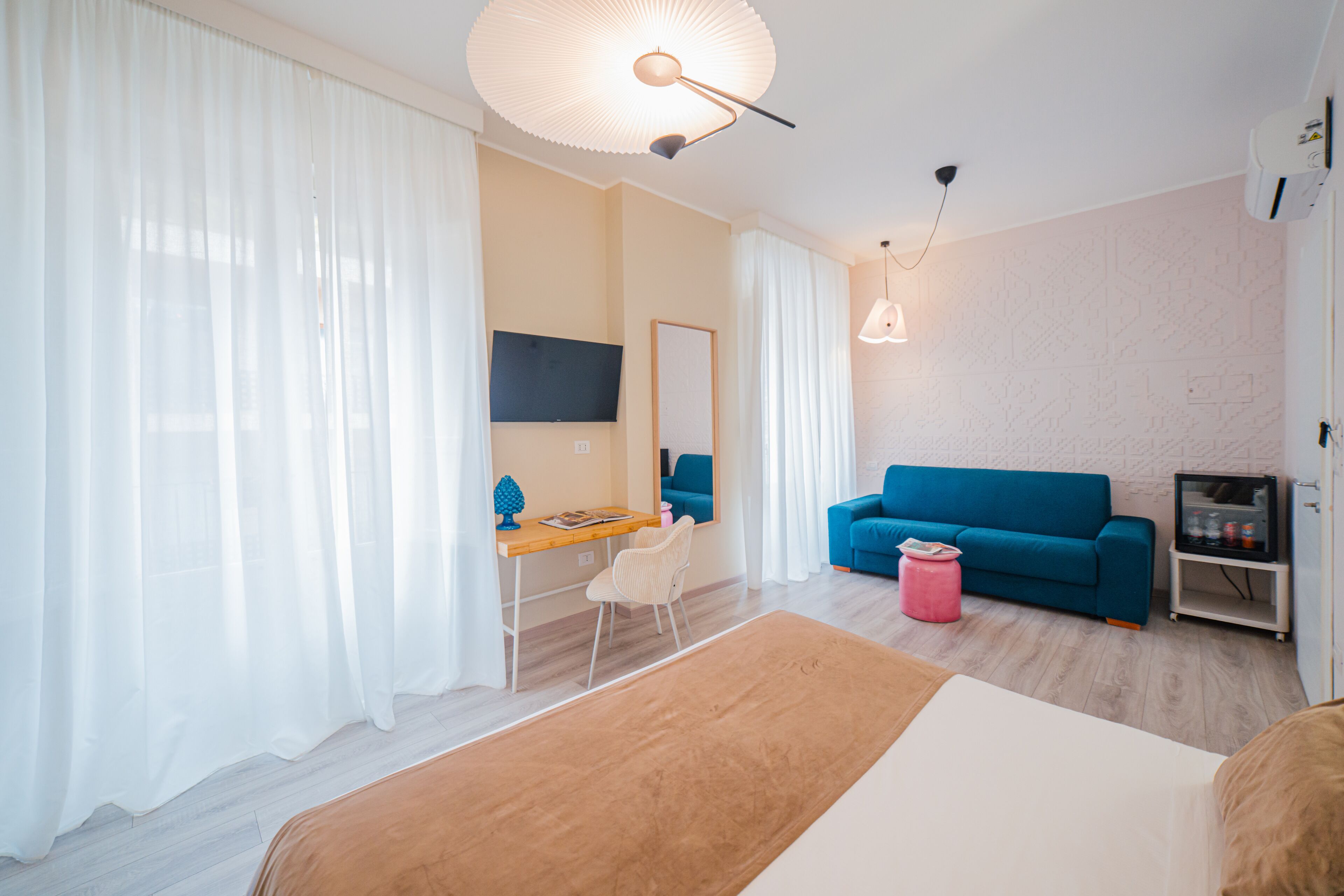 A modern hotel room with a wooden desk, a blue sofa, a bed, and a TV mounted on the wall.