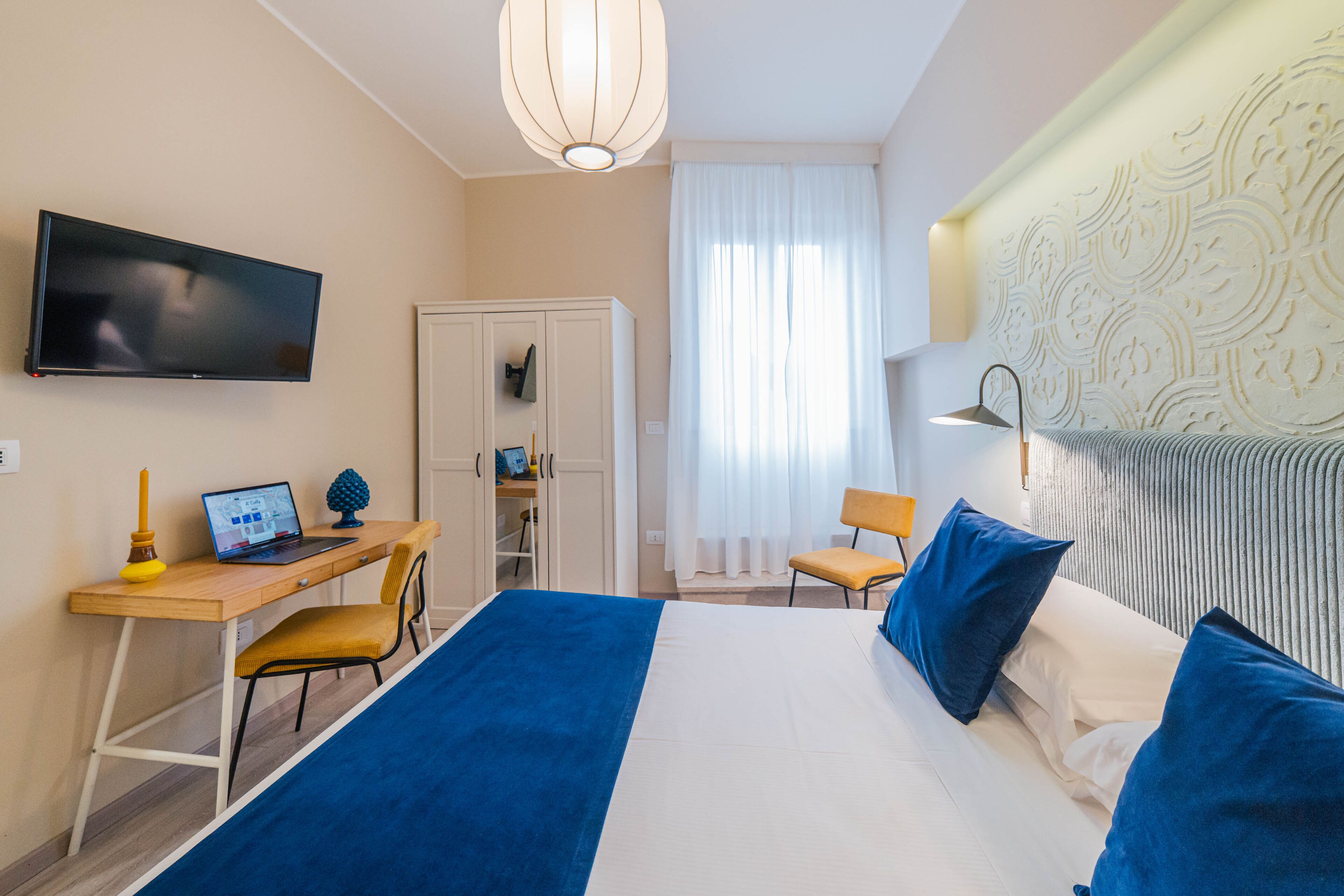 A hotel room with a bed, a desk with a laptop, a chair, a TV, and a window with curtains.