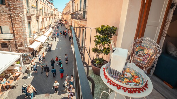 A narrow street lined with buildings, a balcony with a chair and potted plant, and outdoor seating. - A' Coffa (Taormina)