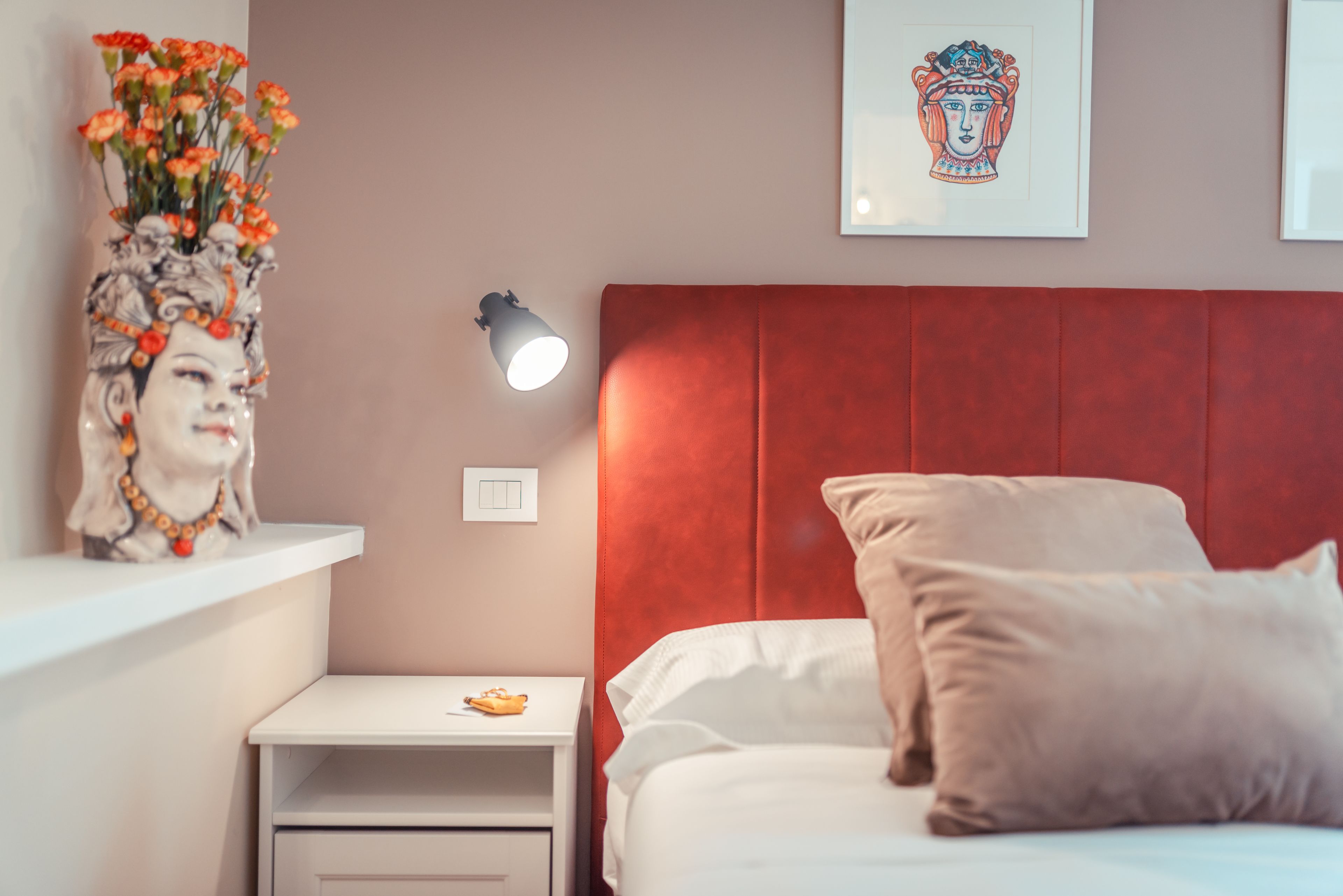 Ein Schlafzimmer mit rotem Kopfteil, einem weißen Nachttisch mit Lampe, einem gerahmten Kunstwerk über dem Bett und einer dekorativen Vase mit Blumen auf dem Nachttisch.