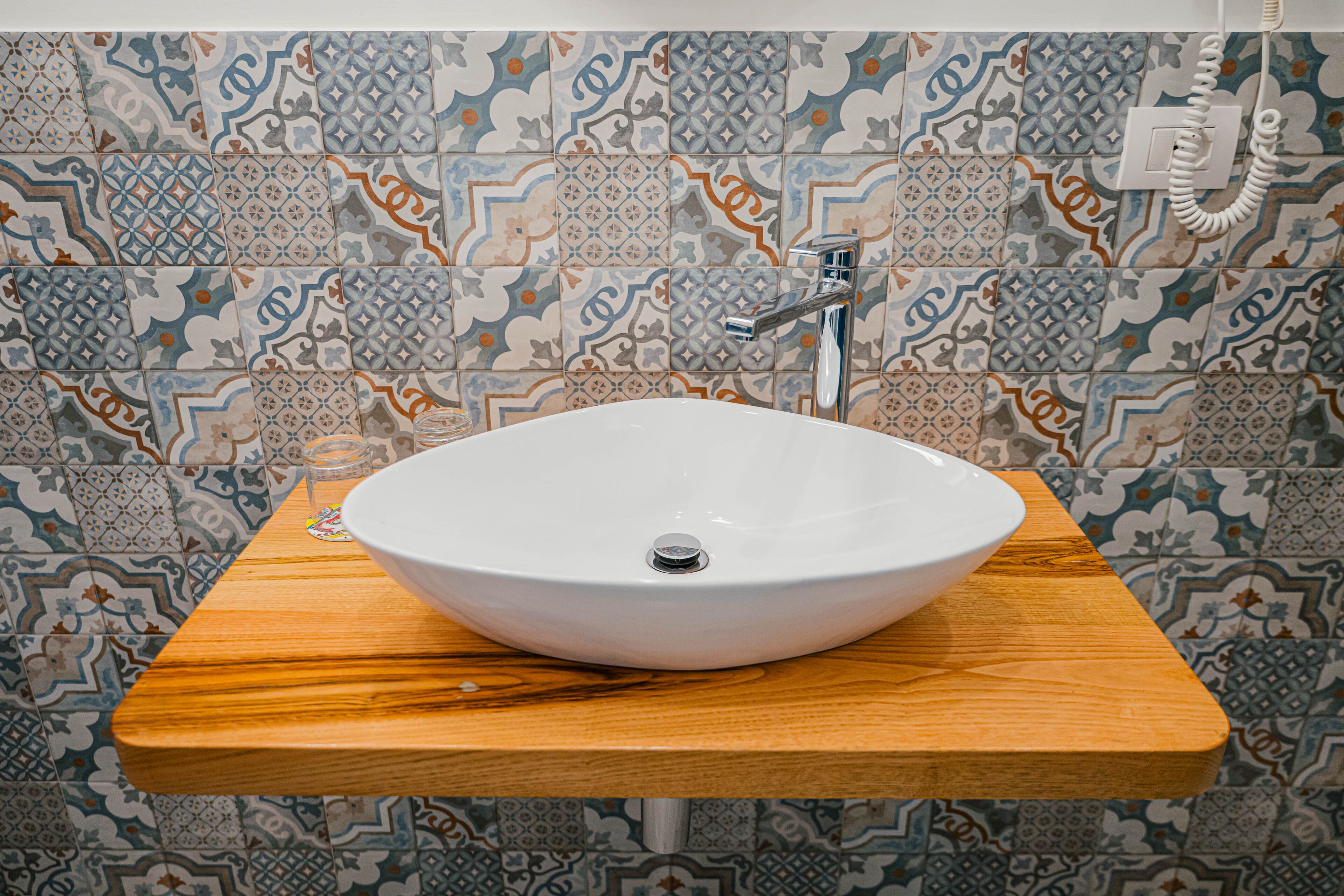 A bathroom with a white sink on a wooden countertop, patterned tiles on the walls, and a visible electrical outlet.