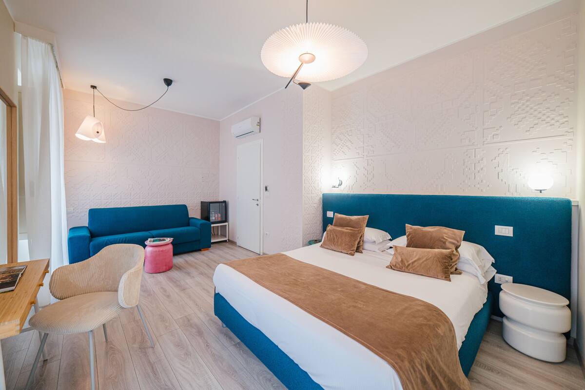 A modern hotel room with a large bed, a blue headboard, a blue sofa, a wooden table, and a chair.