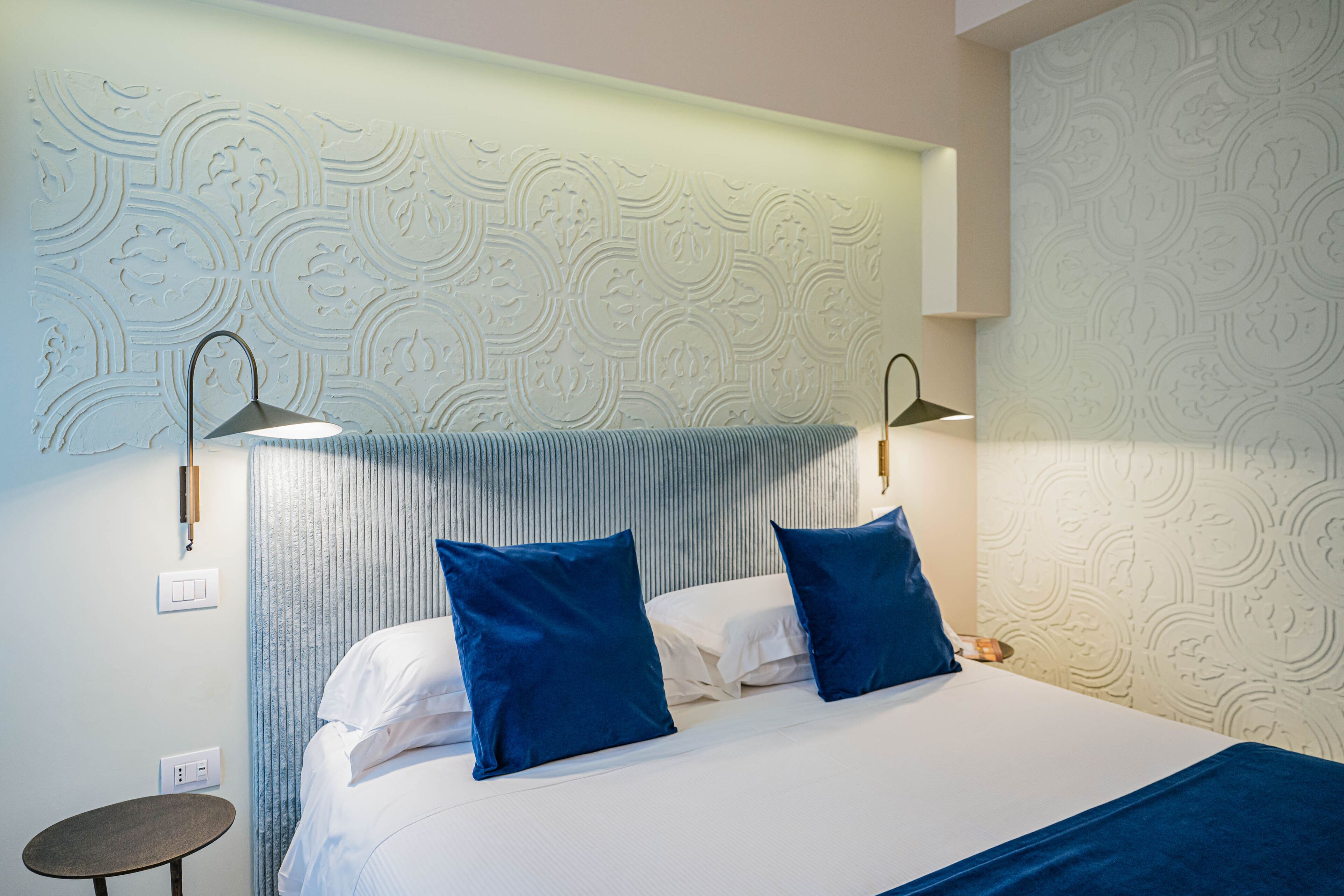 A hotel room with a bed, two blue pillows, a bedside table, and a wall with a decorative pattern.