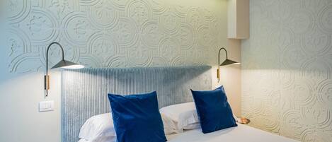 A hotel room with a bed, two blue pillows, a bedside table, and a wall with a decorative pattern.