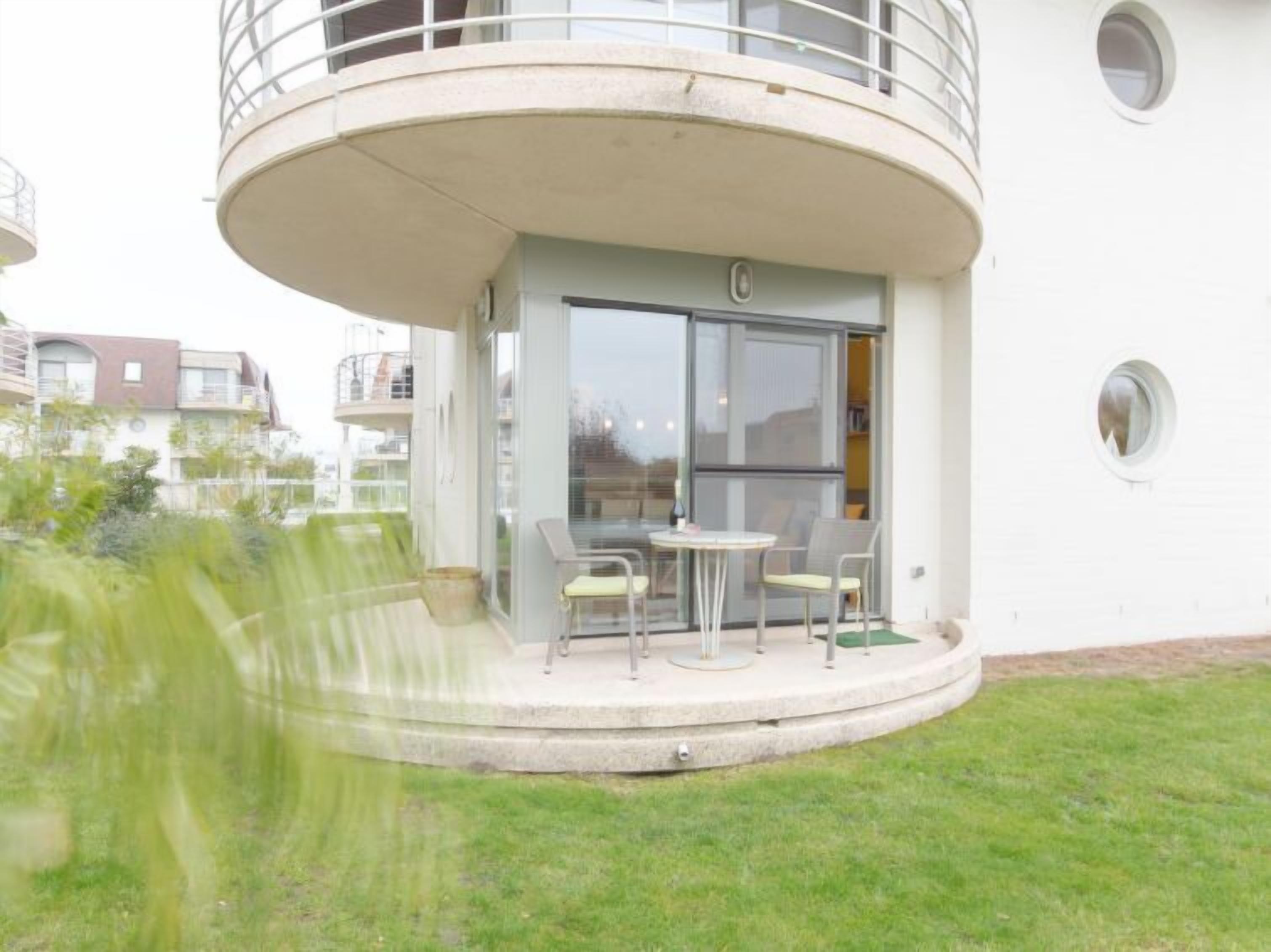 Ein modernes Gebäude mit einem runden Balkon, einem Glastür-Eingang und einer kleinen Außenbereich-Sitzgelegenheit mit Stühlen.