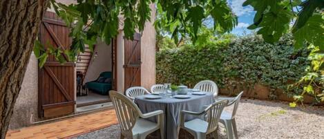 Un espacio para comer al aire libre con una mesa redonda y sillas, rodeado de grava, bajo un árbol.