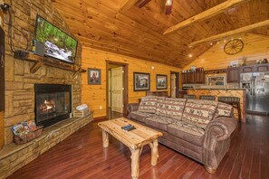 A cozy living room with a stone fireplace, wooden ceiling, and a television mounted on the wall.