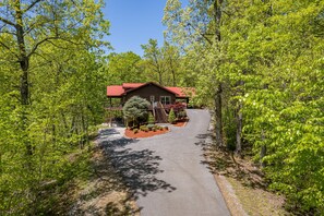 Un chalet avec un toit rouge, entouré d’une végétation luxuriante et verdoyante, et une allée pavée menant à l’entrée.