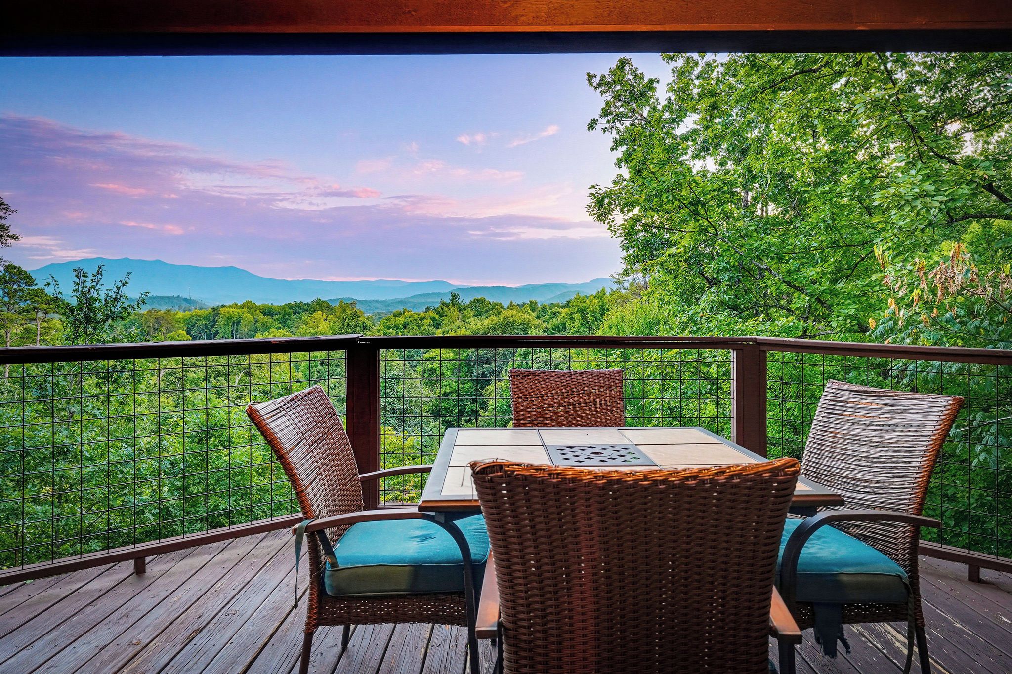 A wooden deck with a table and four wicker chairs, overlooking a lush green landscape and distant mountains.