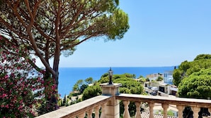 A balcony with a stone balustrade overlooking a coastal landscape with trees and a clear view of the sea.