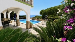 A pool area with a white building, outdoor seating, and a view of the sea.