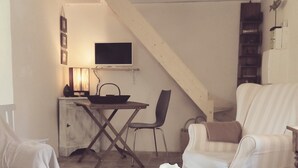 Un salon chaleureux avec un canapé blanc, un fauteuil à rayures et une table basse sur laquelle est posée une bougie. Un escalier mène à une salle à manger équipée d’un téléviseur et d’une lampe.