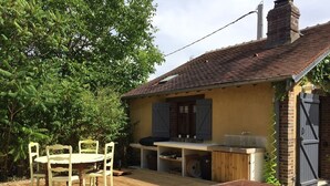 Une terrasse en bois avec une table et des chaises, un banc en bois et un petit espace cuisine extérieur équipé d’un évier et de rangements.