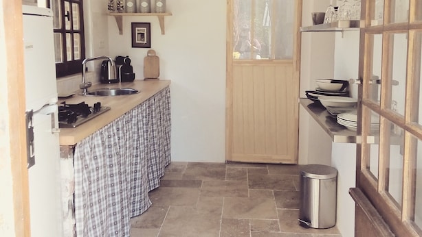 Une cuisine de taille réduite, avec un comptoir en bois, un évier et une cuisinière. On y trouve une poubelle et une porte qui donne sur une autre pièce.