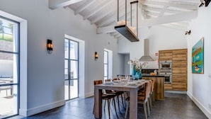 A spacious dining area with a long wooden table and chairs, a kitchen with wooden cabinets, and large windows allowing natural light. - Casa Magnolia - Luxury villa with private heated pool and sea views! (Carvoeiro)
