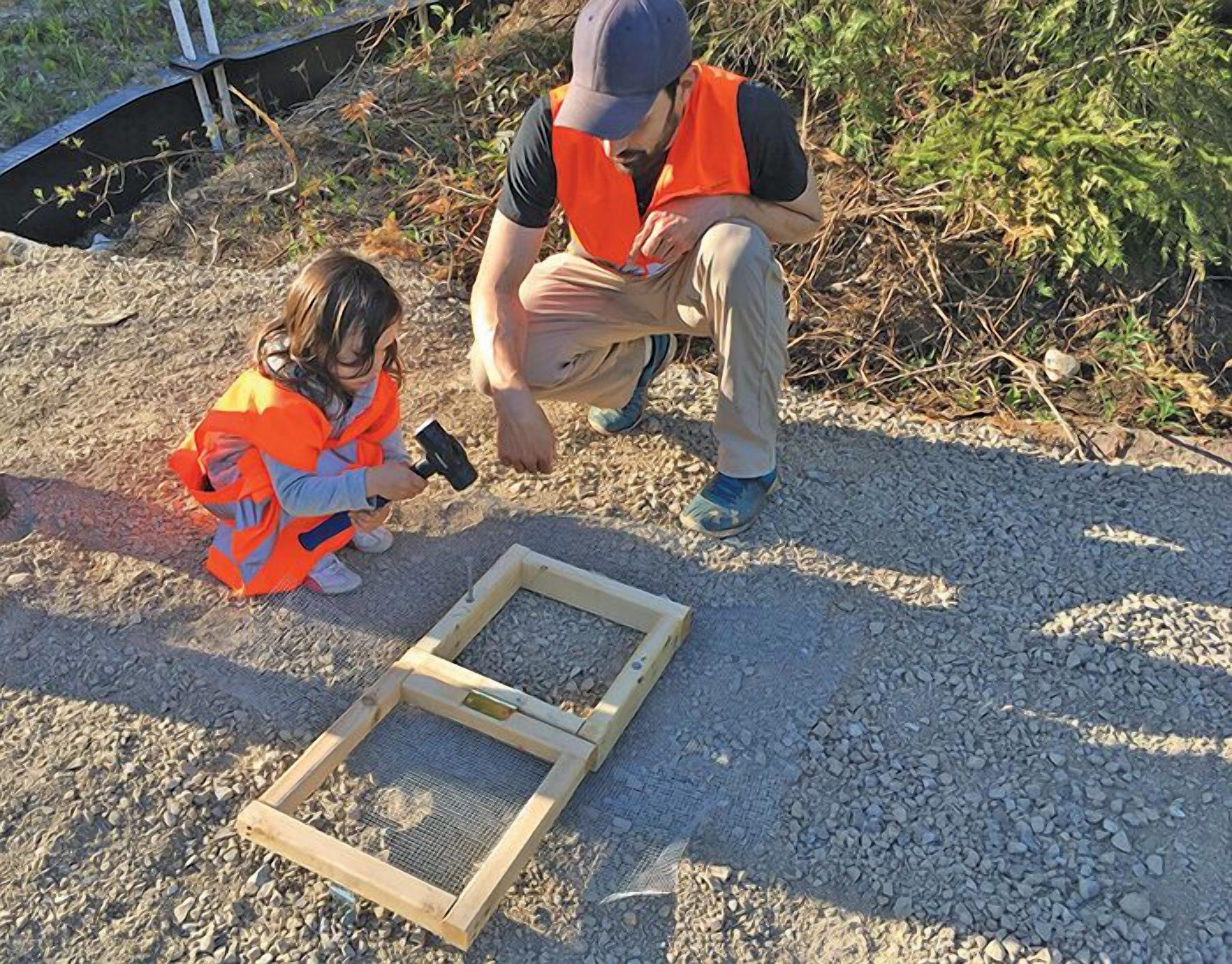 Una persona y un niño observando una estructura de madera sobre un lecho de grava.