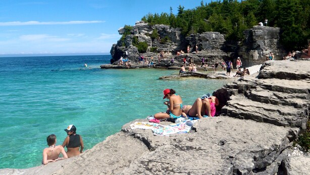 Une côte rocheuse bordée d’une eau turquoise cristalline, des gens se détendant sur les rochers et, en arrière-plan, une falaise couverte de forêt.