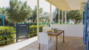 A covered outdoor area with a table and chairs, a view of greenery and trees, and a blue structure in the background.