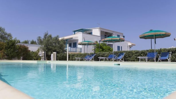 A clear swimming pool with lounge chairs and umbrellas, surrounded by a building with a white facade and blue accents.