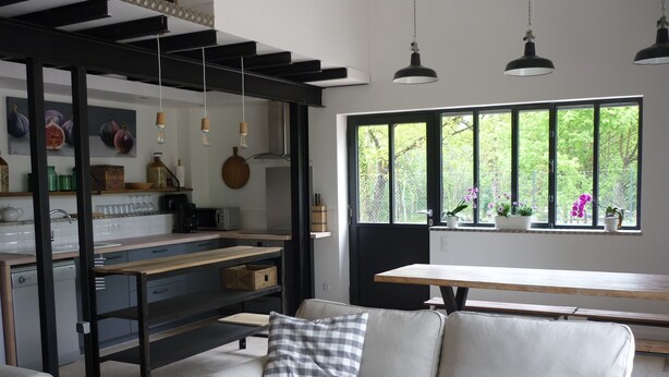 A modern kitchen with a dining area, featuring a sofa with a checkered pillow, pendant lights, and a large window with a view of greenery. - Workshop n°2 for 6 with shared pool and sauna for lazy days (Semur-en-Auxois)