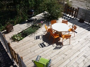 Una terraza de madera con una mesa redonda y sillas anaranjadas, rodeada por una valla metálica y vegetación.
