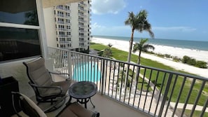 A balcony with a view of the beach, a pool, and a building in the background.